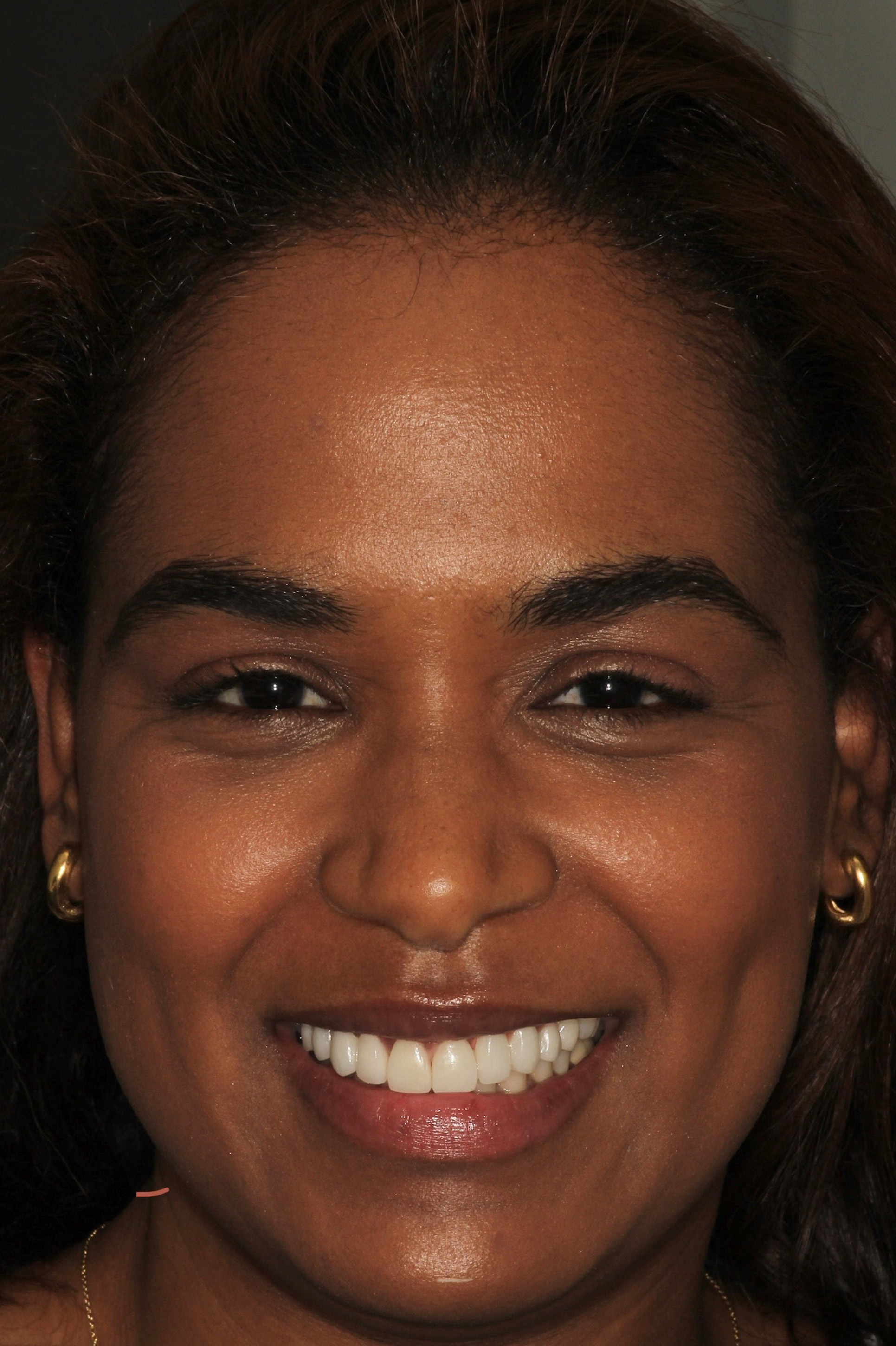 A smiling woman with dark hair and gold jewelry against a neutral background.