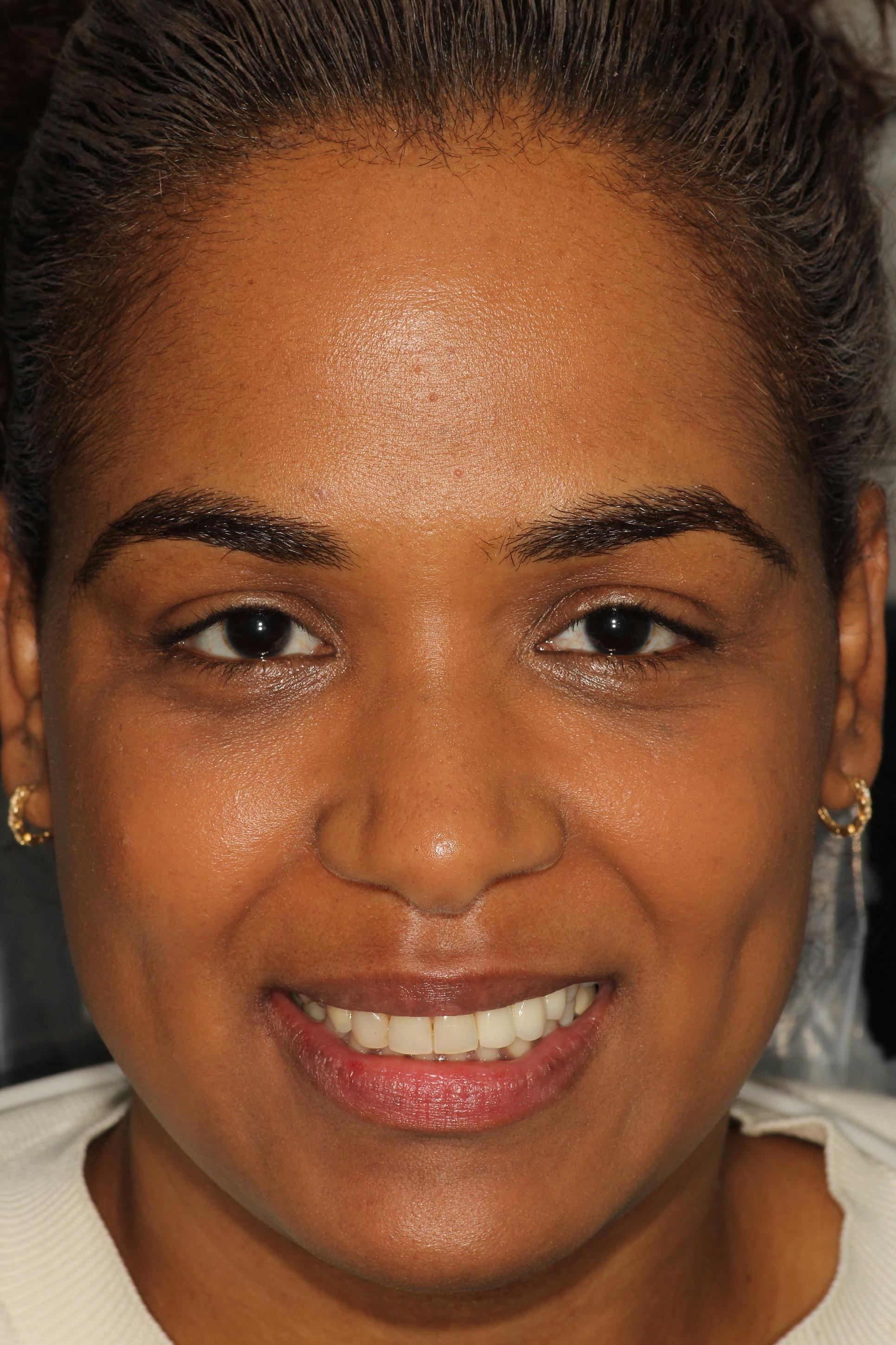 The image shows a smiling woman with dark hair, wearing makeup and a white top, against a plain background.