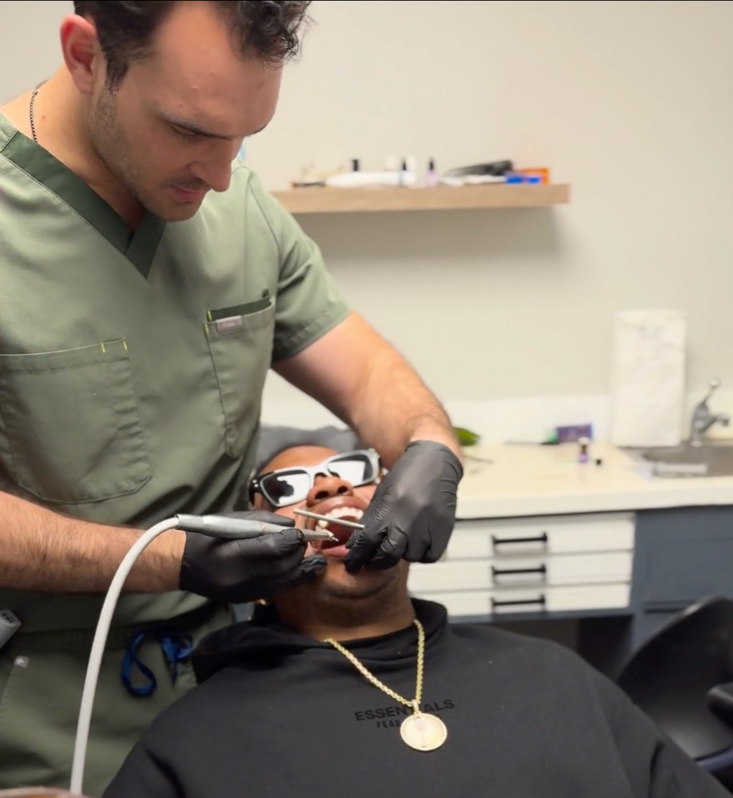 A man wearing sunglasses receives dental care from a professional with a stethoscope around his neck, both seated in an office setting.