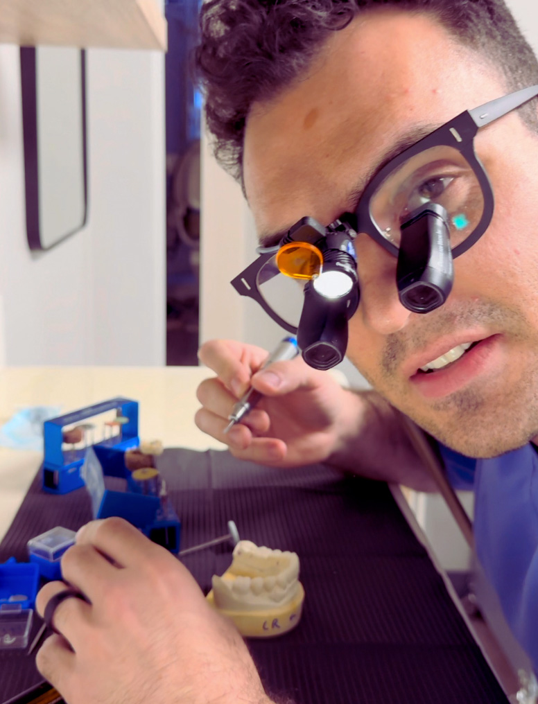 A man wearing glasses is closely examining a small object with magnifying glasses, possibly working on a project or repairing something at a workbench.
