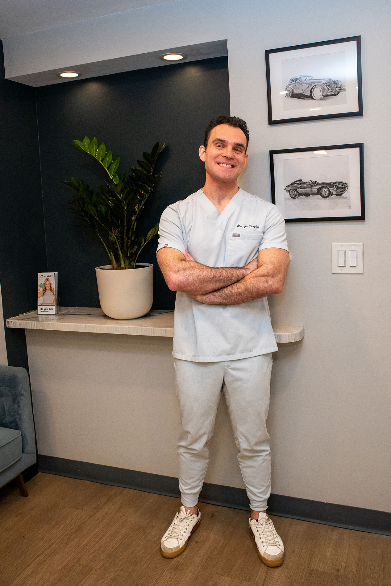 A man wearing a white lab coat stands confidently with his arms crossed in front of him, smiling at the camera, posing for a photo inside an office setting with framed pictures on the wall behind him.