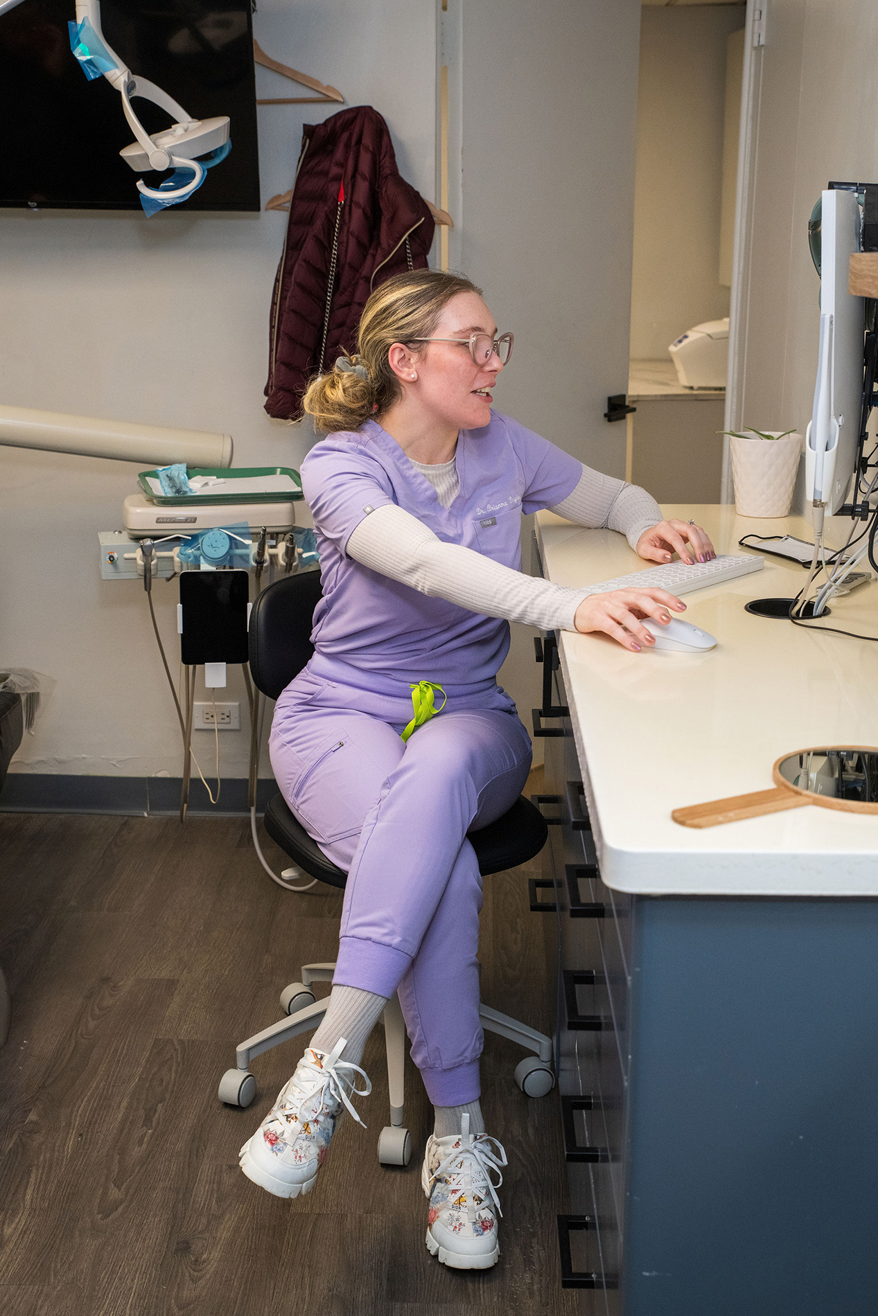 Woman in purple scrubs sitting at desk with computer mouse and keyboard, wearing glasses and headphones.