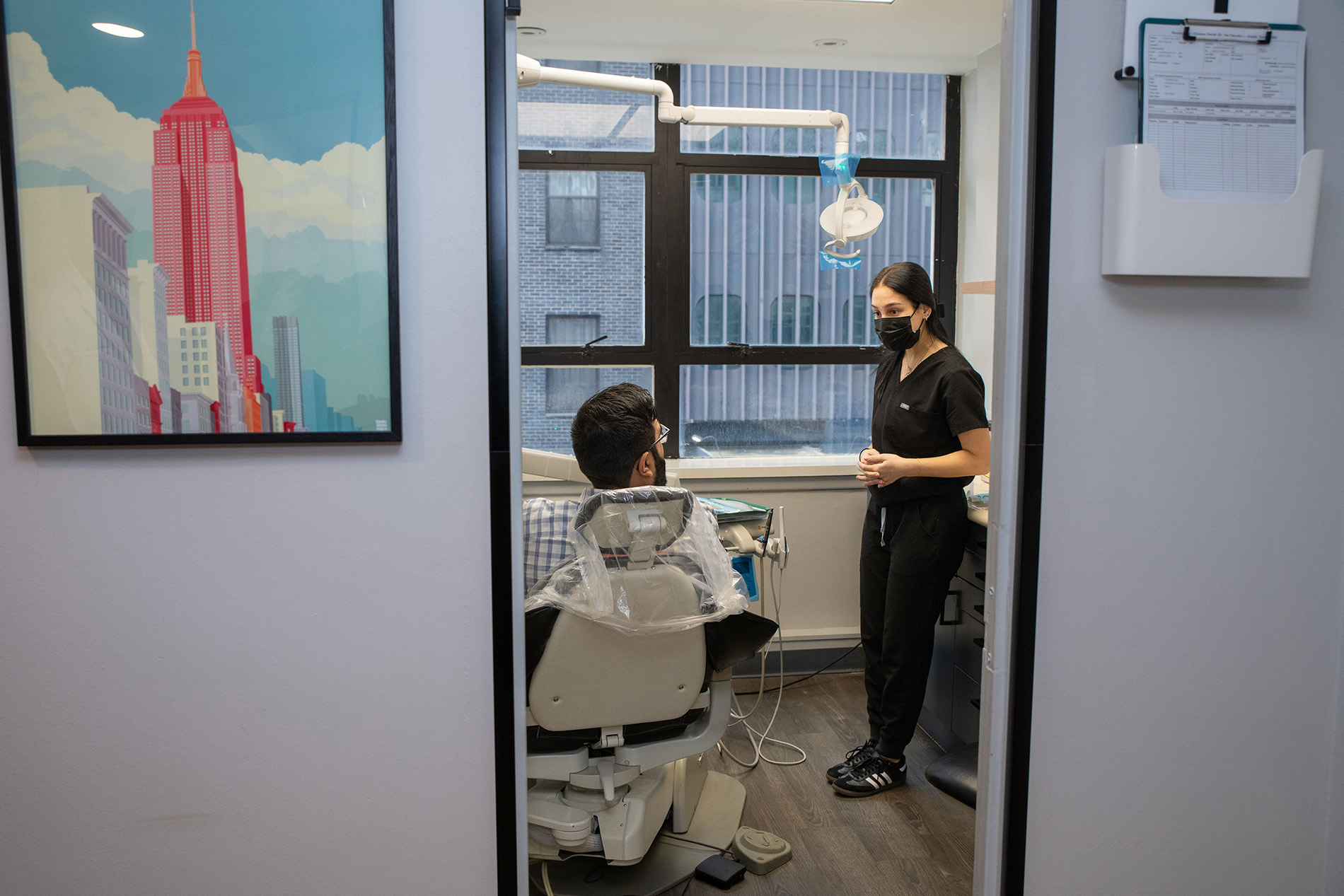 In the image, there is a dental office setting with a dentist standing next to a patient in a chair, both wearing masks, with various dental equipment visible in the background, including a sink, mirror, and posters on the wall featuring cityscapes.