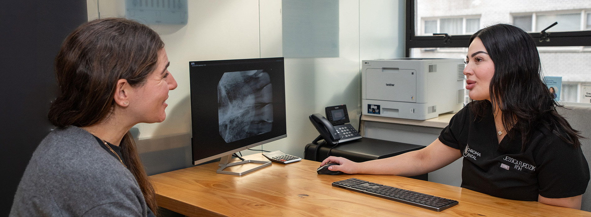 The image shows two people sitting at a desk with a computer monitor between them, where one person appears to be speaking to the other.